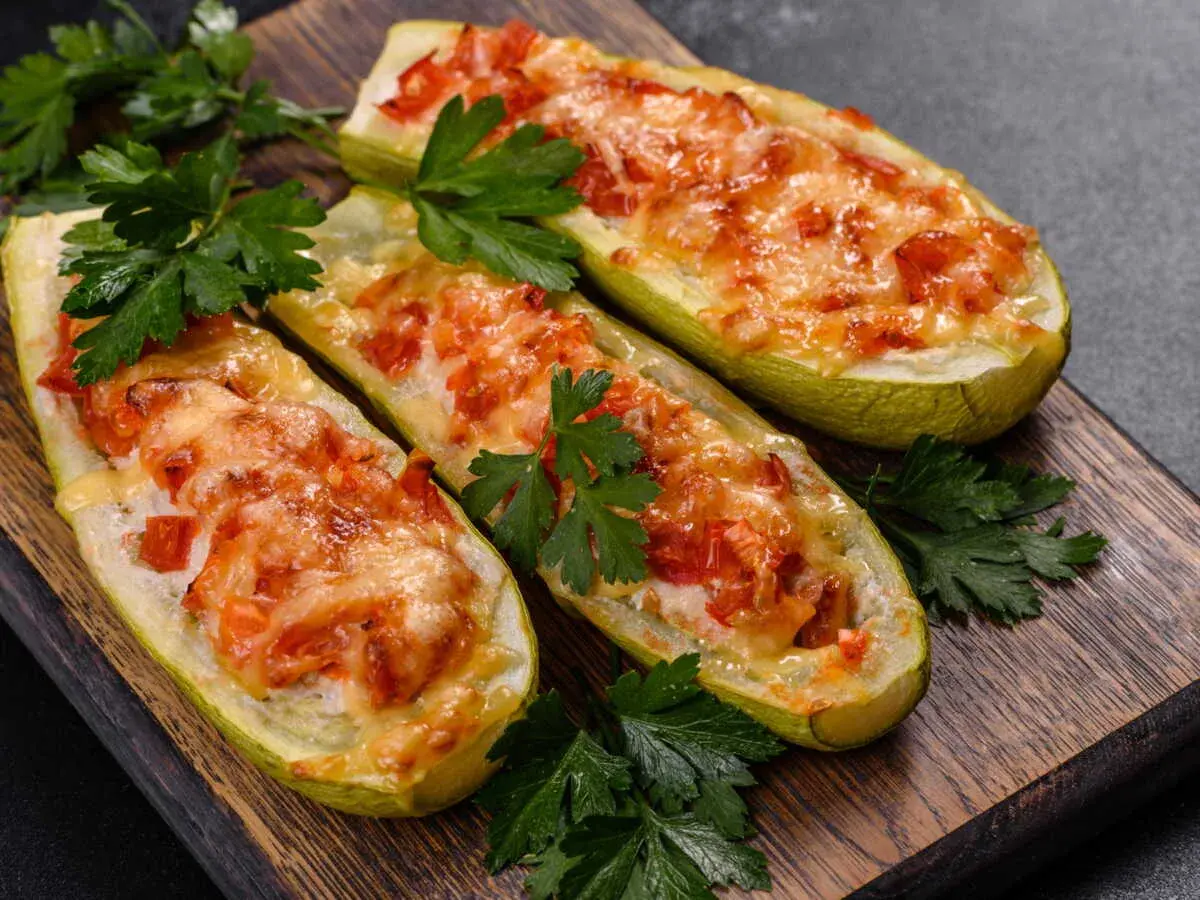 Trois courgettes farcies, façon pizza, gratinées au fromage et garnies de tomates, sur une planche en bois, décorées de persil frais.