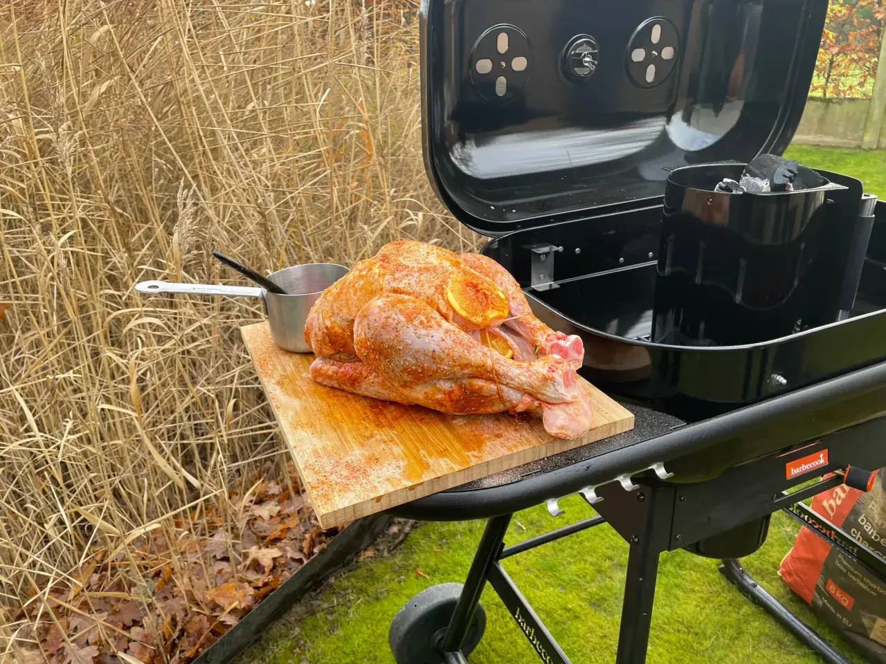 Un dindon assaisonné prêt pour un barbecue de Noël, posé sur une planche à découper à côté d'un barbecue ouvert.