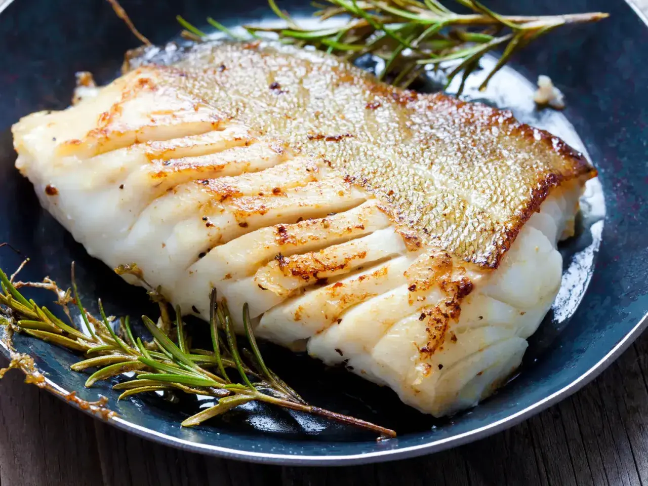 Un filet de poisson blanc parfaitement cuit, peau dorée et chair nacrée, servi avec du romarin frais. Une belle illustration de comment cuire du poisson blanc.