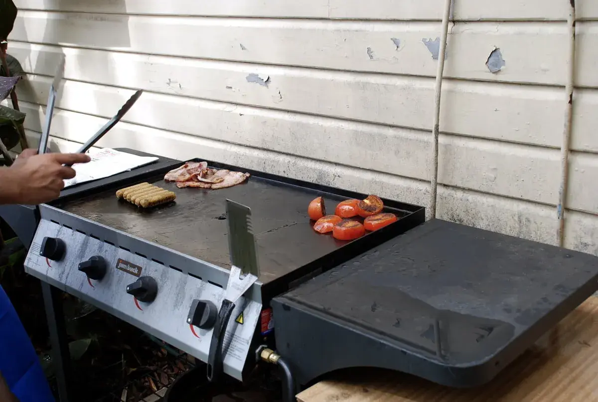 Un barbecue avec une bouteille de gaz prépare un petit-déjeuner savoureux : saucisses, bacon et tomates grillées.