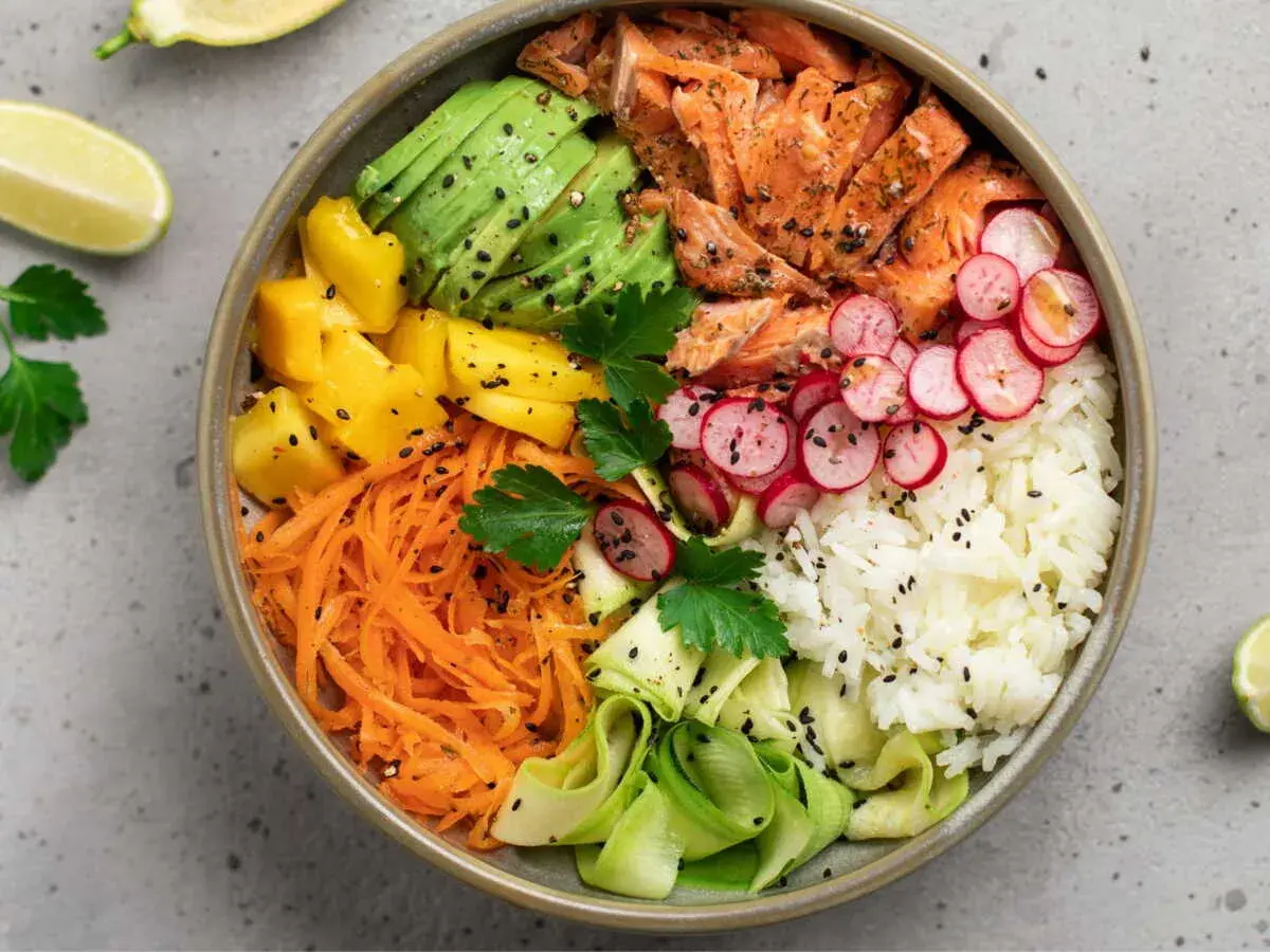 Un bol coloré avec saumon, avocat, mangue, carottes râpées, radis, courgette et riz blanc. Une recette parfaite pour un repas léger en juillet.
