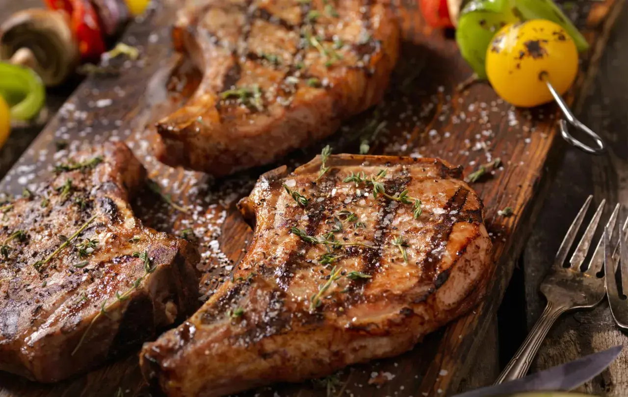 Trois côtelettes de porc grillées, garnies d'herbes et de gros sel, sur une planche en bois. Une surprise de porc barbecue prête à être dégustée.