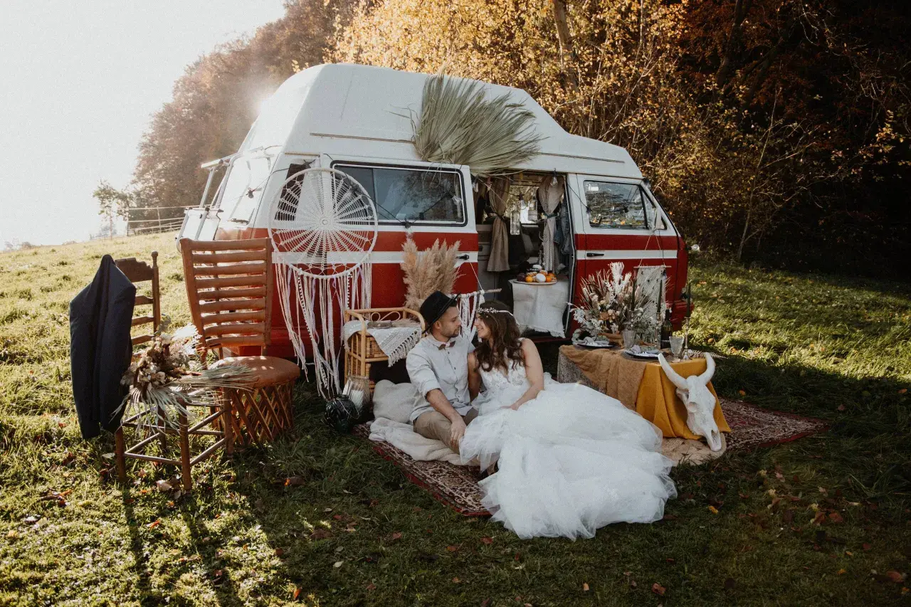 Un couple en tenue de mariage profite d'un mariage pique-nique champêtre près d'un van aménagé, décoré de style bohème.