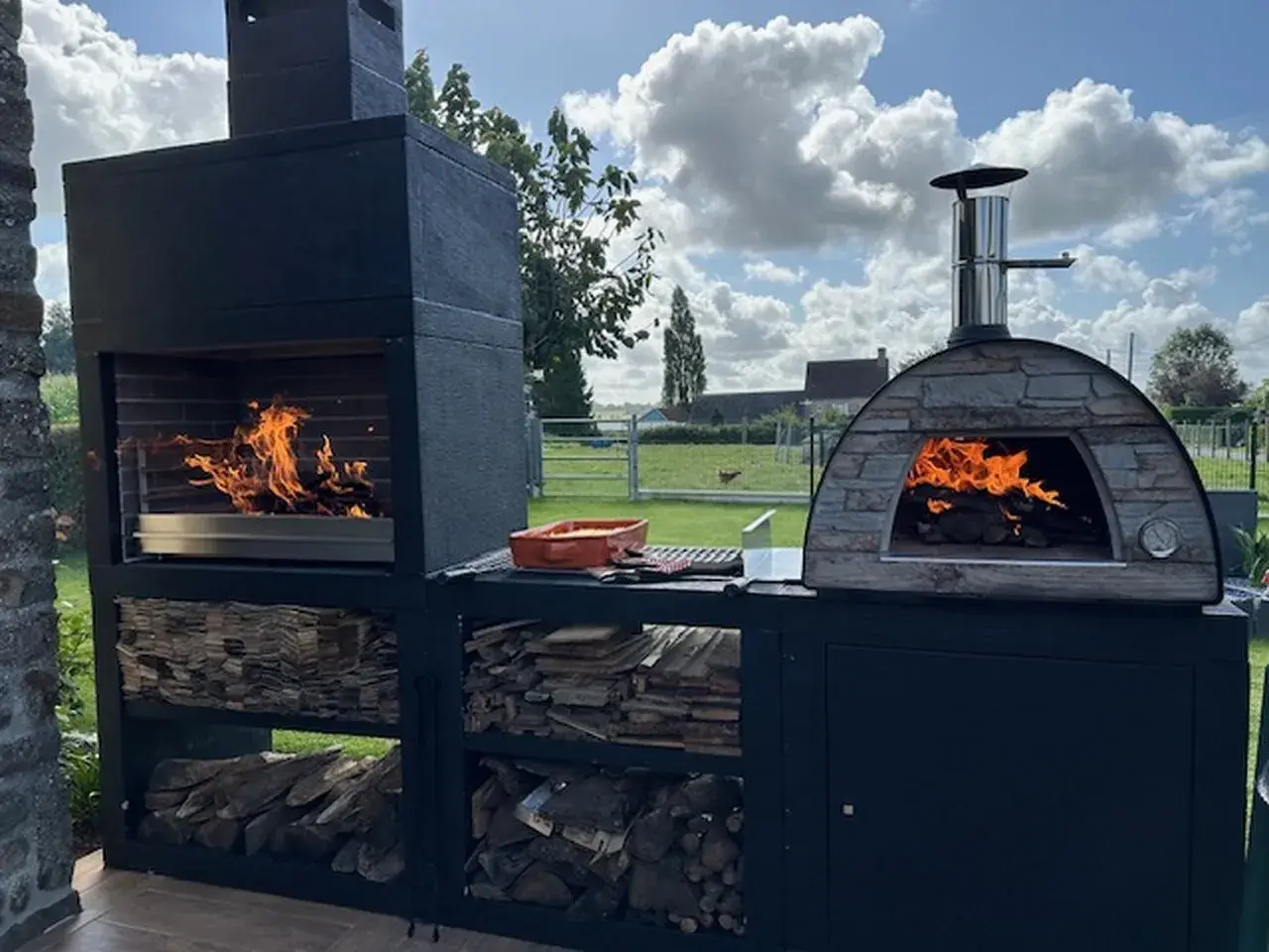 Un barbecue et un four à pizza en pierre, tous deux allumés, prêts pour un repas. Idéal pour savoir comment faire un barbecue sans charbon, avec du bois stocké en dessous.