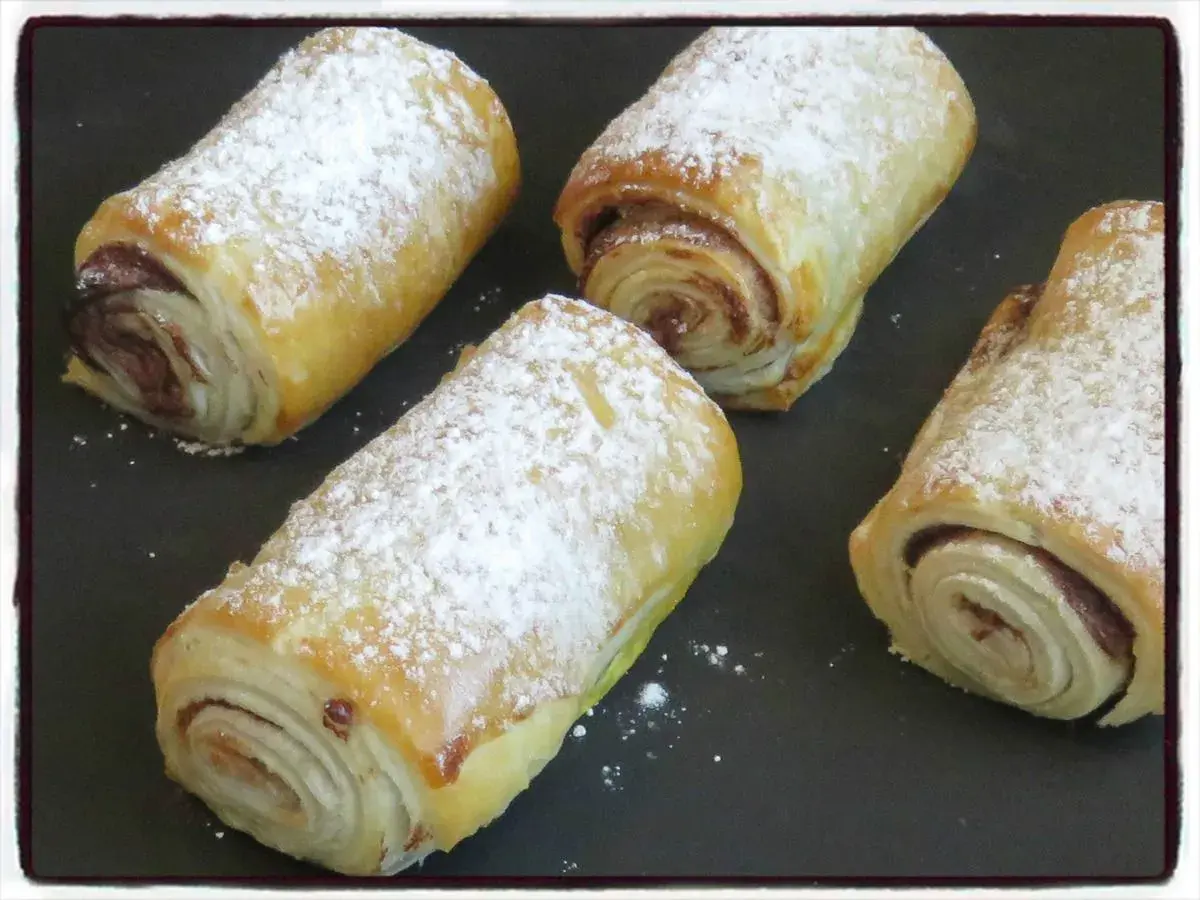 Quatre croissants au chocolat en pâte feuilletée, saupoudrés de sucre glace, prêts à être dégustés.