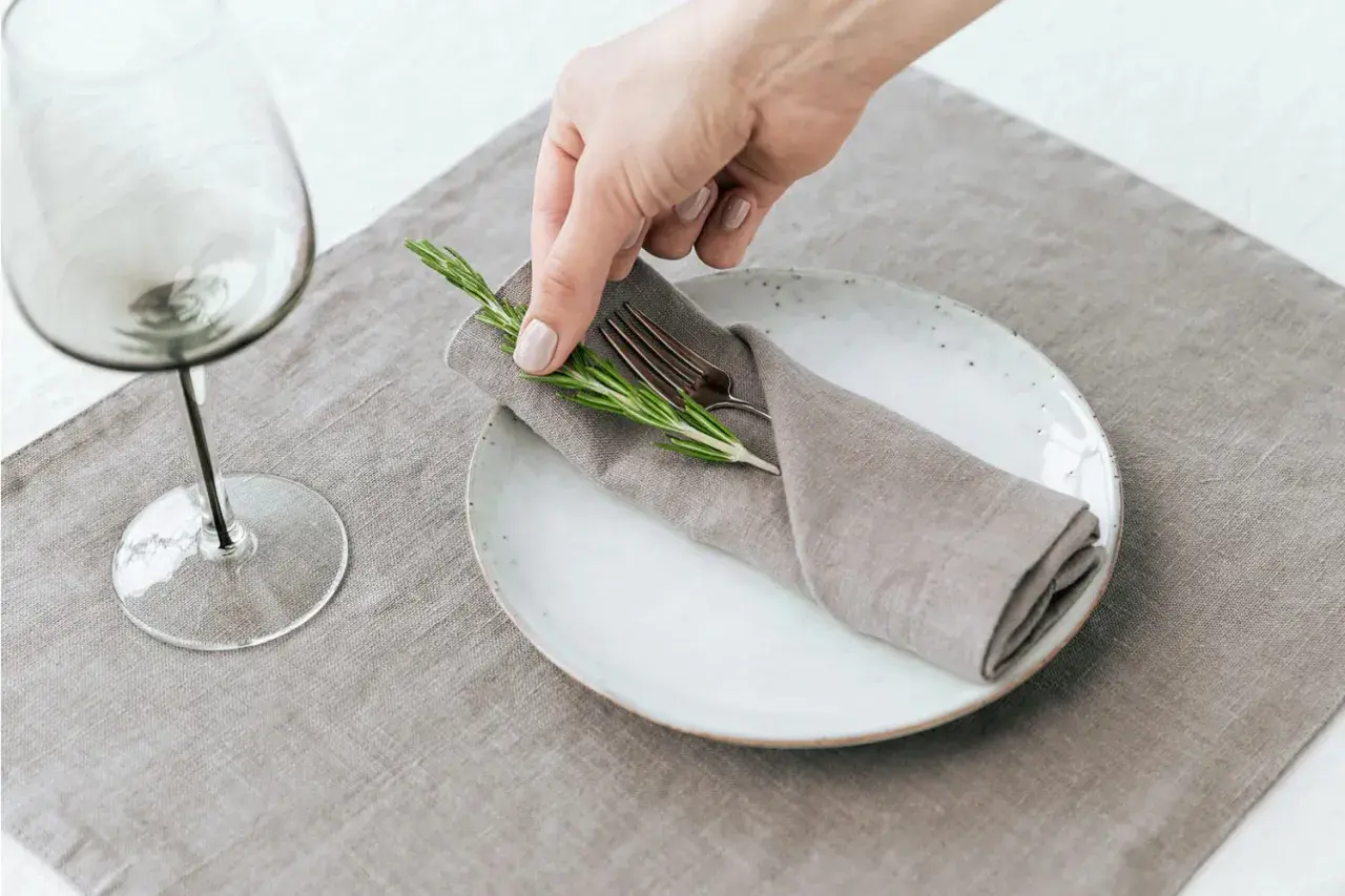 Une main dépose une branche de romarin sur un pliage de serviette mariage, sur une assiette blanche.