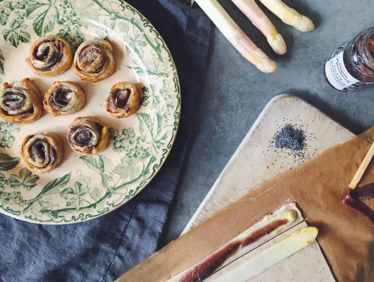 Petits roulés feuilletés à l'asperge blanche, parfaits pour un apéritif. Des asperges blanches, des graines de pavot et une touche de anchois complètent la scène.
