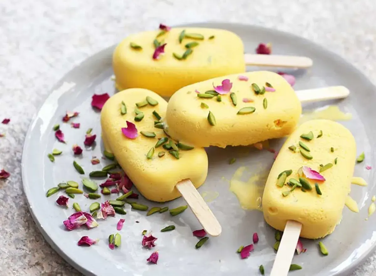 Quatre glaces jaunes, garnies de pistaches et de pétales de rose, sur une assiette. Un délicieux dessert iranien.