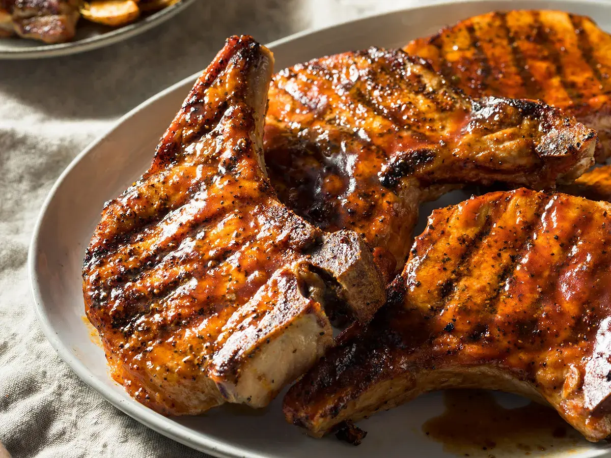 Trois côtes de porcelet grillées, nappées d'une sauce caramélisée et parsemées de poivre noir, sur une assiette blanche.