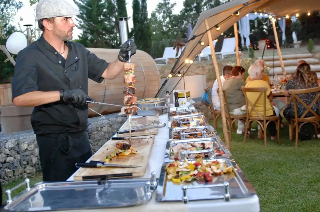 Un chef prépare un menu barbecue mariage. Des brochettes de viande et des légumes sont présentés dans des plats chauffants, tandis que des invités dînent sous une tente décorée de guirlandes lumineuses.
