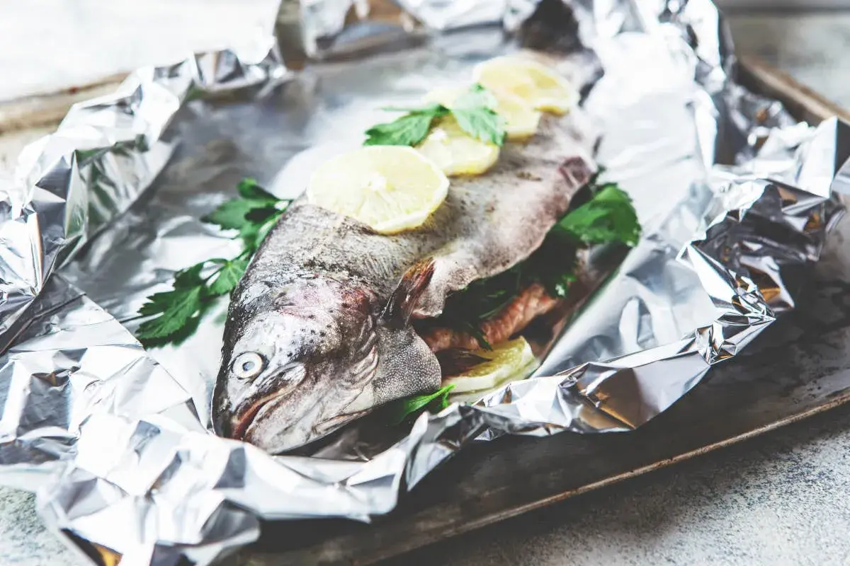 Une truite entière, prête à être cuite en papillote, garnie de rondelles de citron et de persil frais, sur une feuille d'aluminium froissée.
