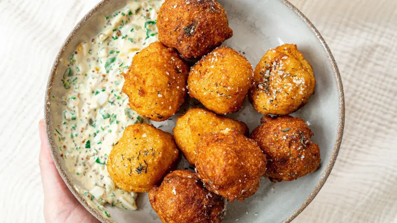 Un bol de pommes dauphines croustillantes, dorées et saupoudrées d'herbes, accompagnées d'une sauce crémeuse aux herbes et aux œufs.