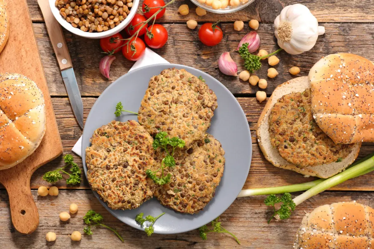 Trois galettes de lentilles, parfaites pour une recette de steak végétarien, sont présentées avec des pains à burger, des tomates et de l'ail sur fond de bois.