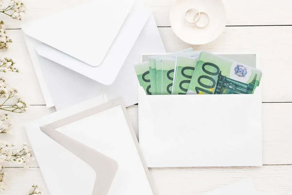 Enveloppes blanches, fleurs et billets de 100€ pour savoir comment offrir de l'argent pour un mariage. Bague de fiançailles sur une soucoupe.