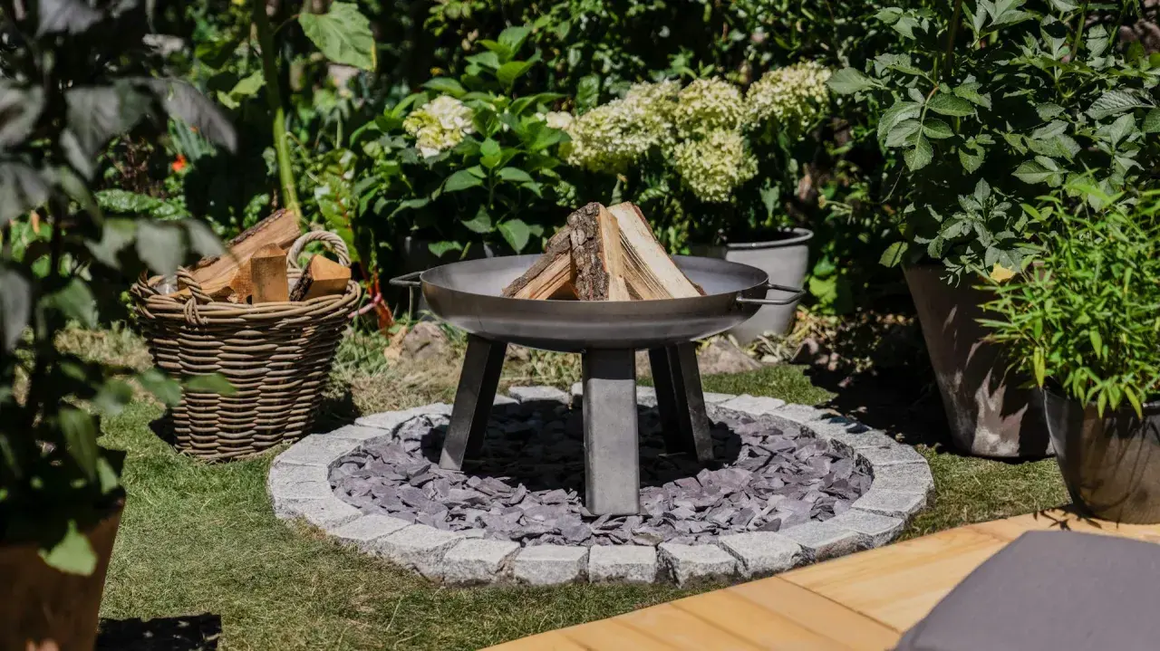 Un brasero en forme de chaudron, prêt à être allumé, avec du bois à côté. Un coin de jardin accueillant pour des soirées conviviales.