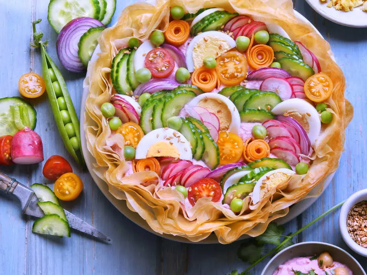 Un plat d'été raffiné : une tarte salée garnie de concombre, radis, œufs durs, tomates cerises, petits pois et oignons rouges, le tout sur un lit de crème rose.
