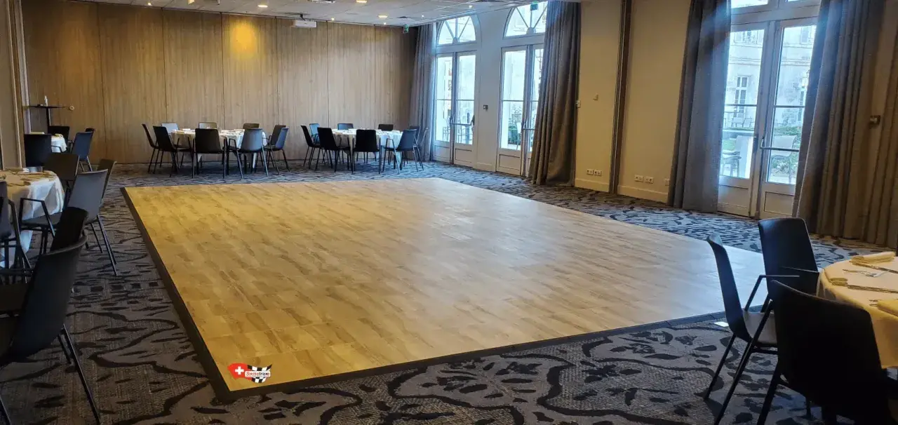 Une piste de danse en bois clair, idéale pour savoir comment faire une piste de danse pas cher. Des tables rondes et des chaises noires sont disposées autour.