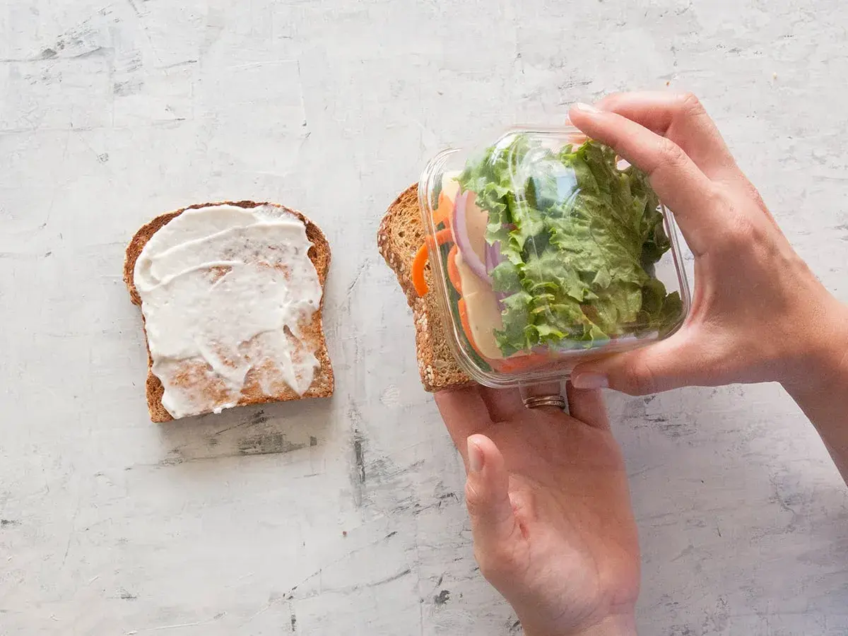 Préparation d'un sandwich crudité : une tranche de pain tartinée de mayonnaise, l'autre garnie de salade, carottes, oignons et fromage, prête à être assemblée.