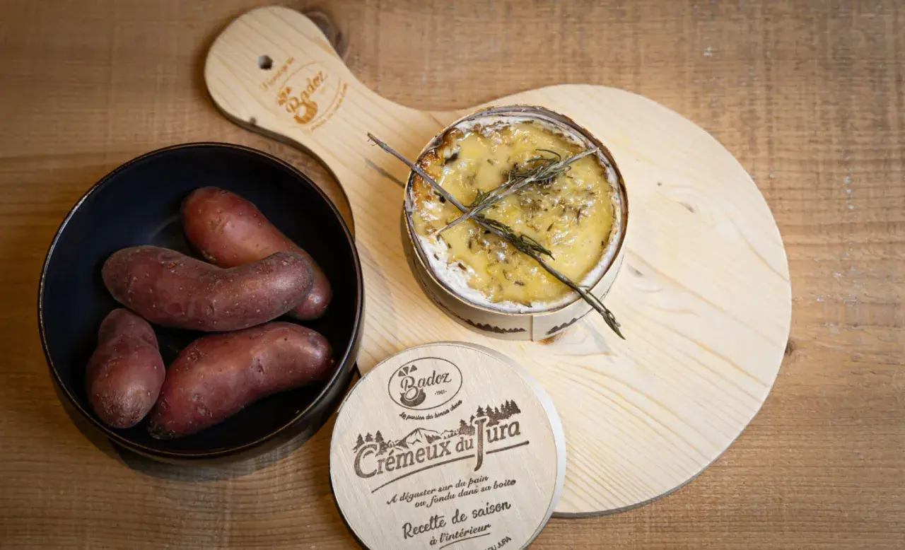 Un crémeux du Jura au four, doré et fondant, avec des pommes de terre rouges dans un bol. Une planche en bois ornée du logo "Crémeux du Jura" complète la scène.