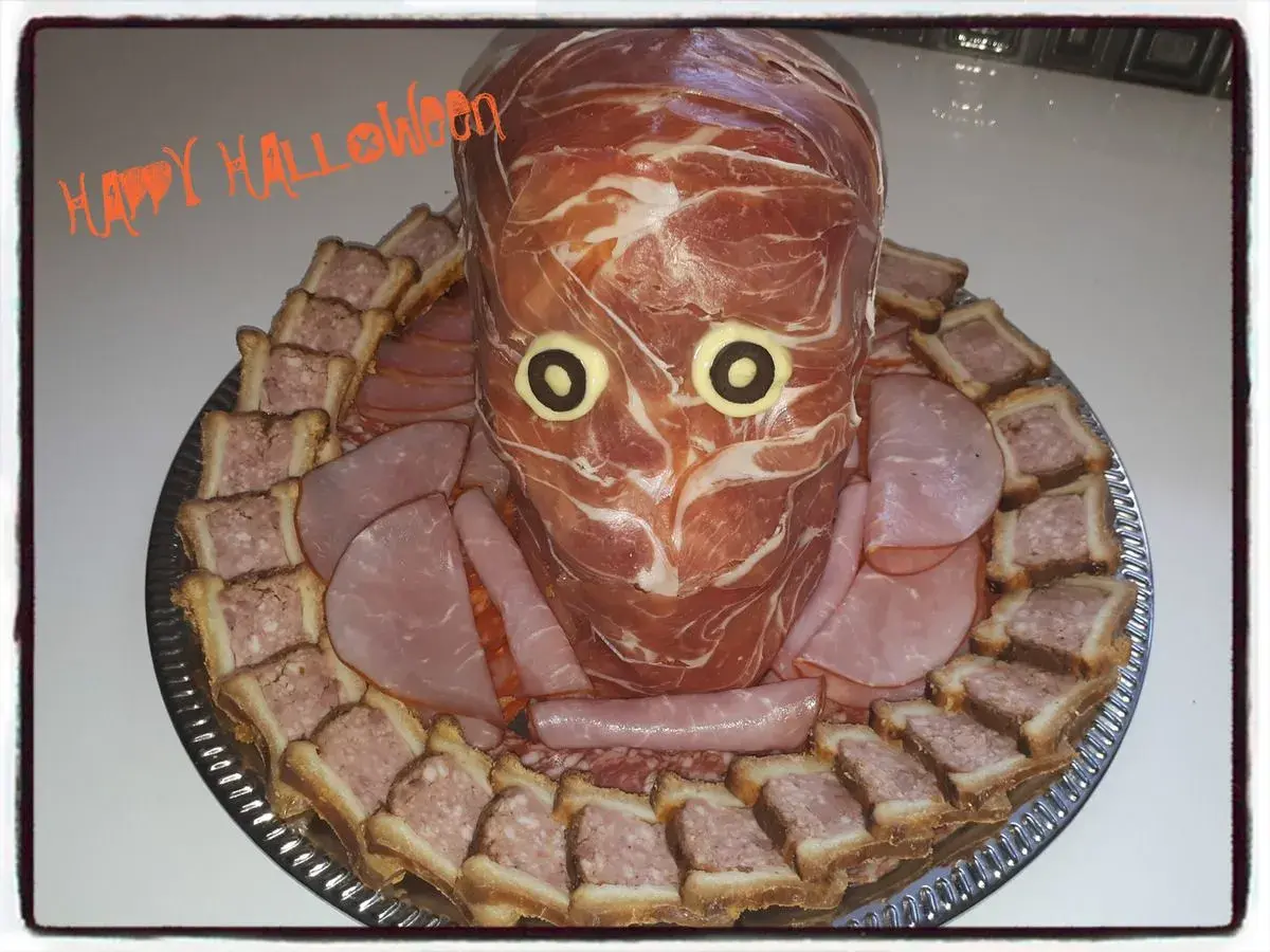 Un plateau de crudités d'Halloween en forme de tête de momie, entouré de petits sandwichs et de charcuterie.