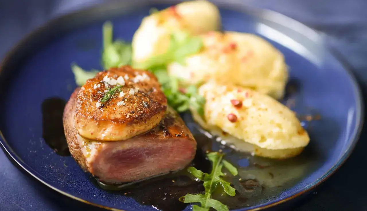 Tournedos de canard cuisson parfaite, servi avec une purée aérienne et des herbes fraîches sur une assiette bleue.