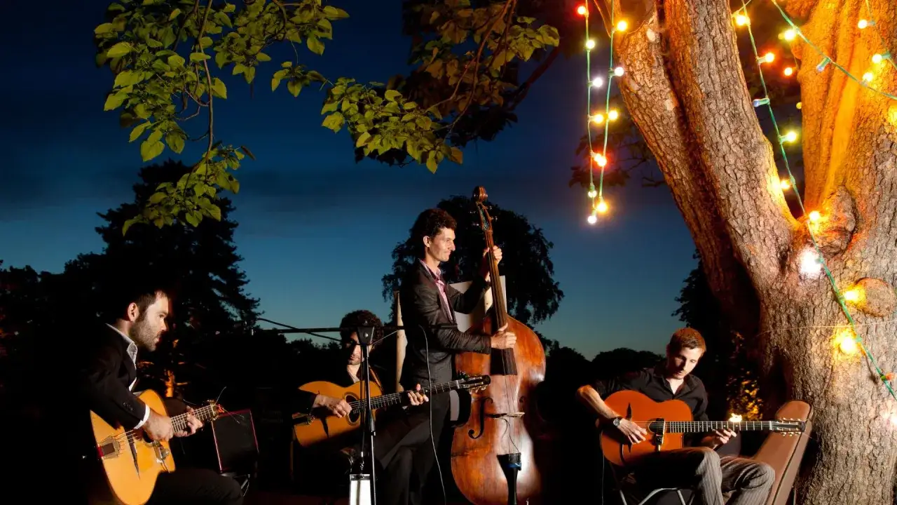 Un groupe de musique joue en extérieur à la nuit tombée, éclairé par des guirlandes lumineuses. Le prix de ce concert est une soirée inoubliable.