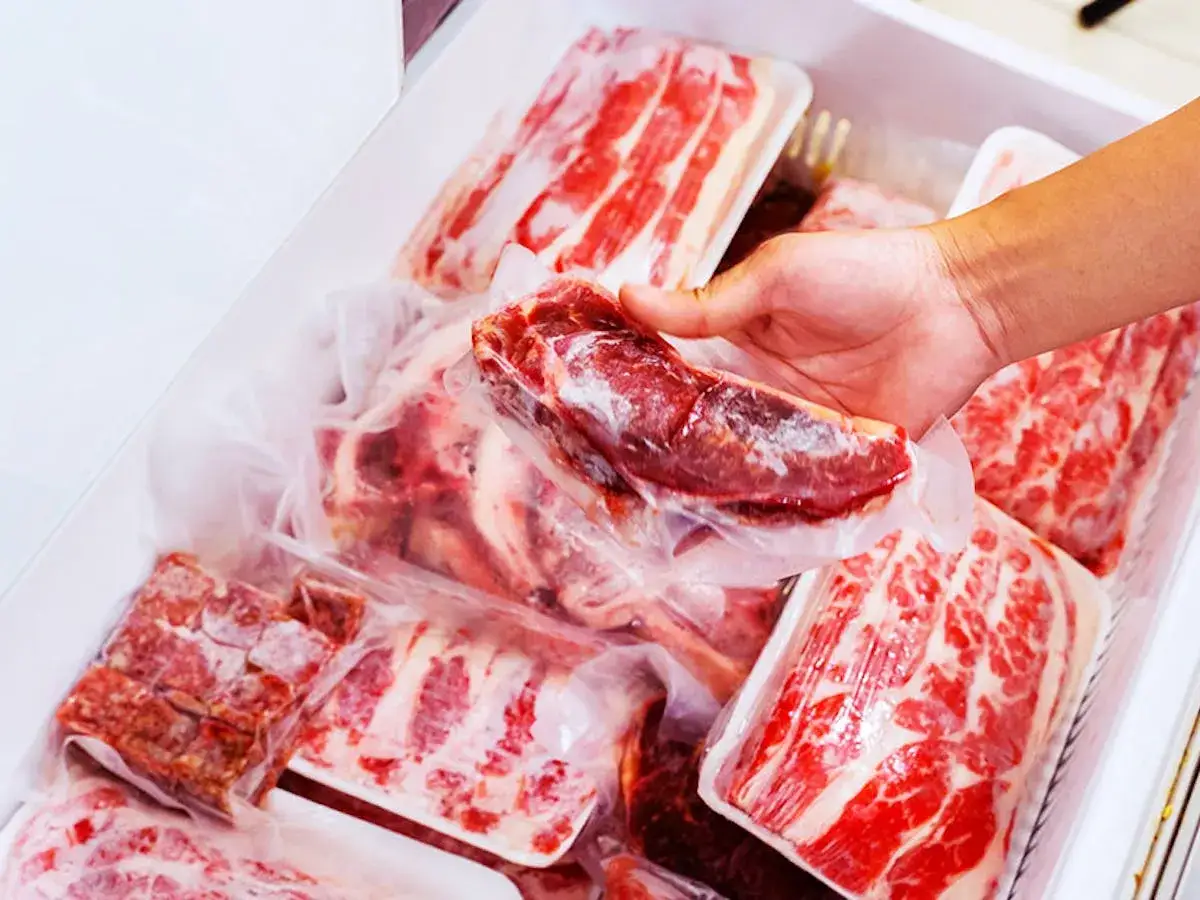 Main d'une personne sortant une pièce de viande sous vide hors du frigo, parmi d'autres paquets de viande.