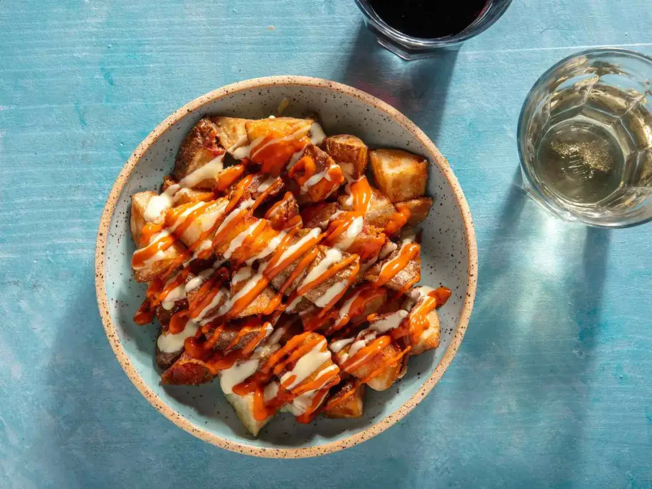 Patatas bravas croustillantes arrosées d'une sauce piquante et d'une sauce crémeuse, servies dans un bol sur une table bleue.