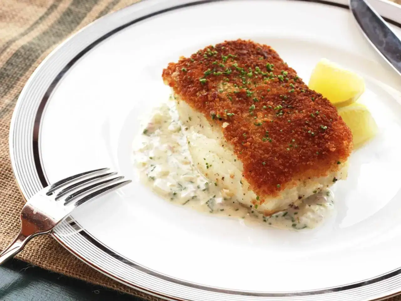 Un filet de poisson blanc pané, servi sur une sauce crémeuse avec des quartiers de citron. Une belle façon de comment cuire du poisson blanc.