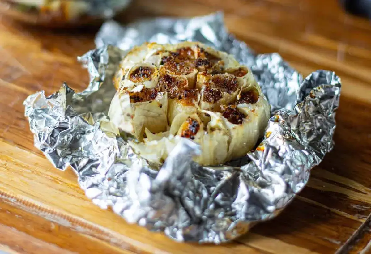 Une tête d'ail au barbecue, dorée et caramélisée, enveloppée dans du papier d'aluminium froissé sur une planche en bois.