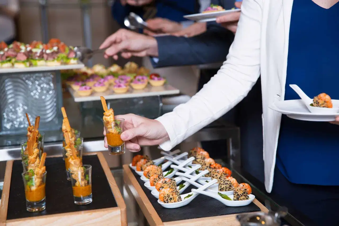 Un buffet repas avec des verrines de crevettes et des brochettes de poulet. Des convives se servent.