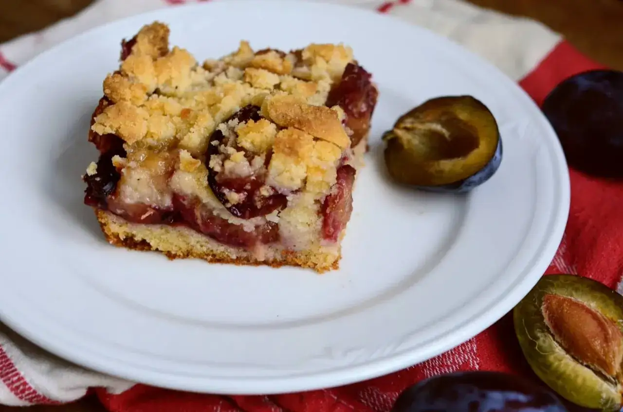 Tranche de g&acirc;teau aux prunes &agrave; la po&ecirc;le, garnie de crumble dor&eacute; et de fruits juteux. Une demi-prune &agrave; c&ocirc;t&eacute;.