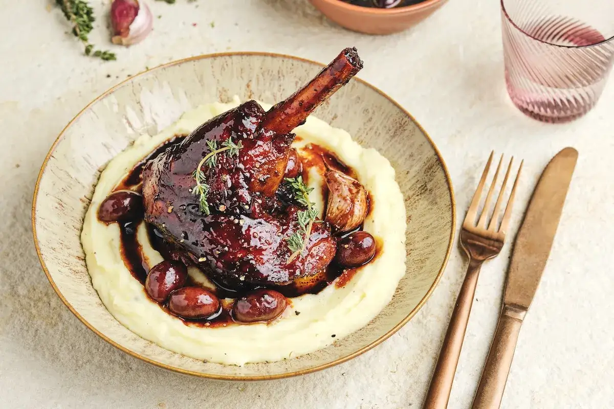 Souris d'agneau au barbecue Weber, nappée d'une sauce foncée, servie sur une purée onctueuse avec des olives et de l'ail rôti.