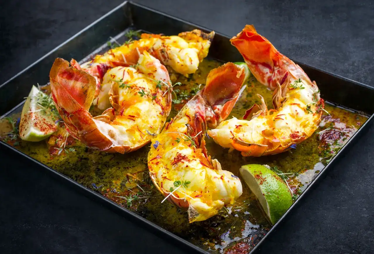 Queue de langouste plancha, dorée et savoureuse, baignant dans une sauce verte, garnie de thym et de quartiers de citron vert.