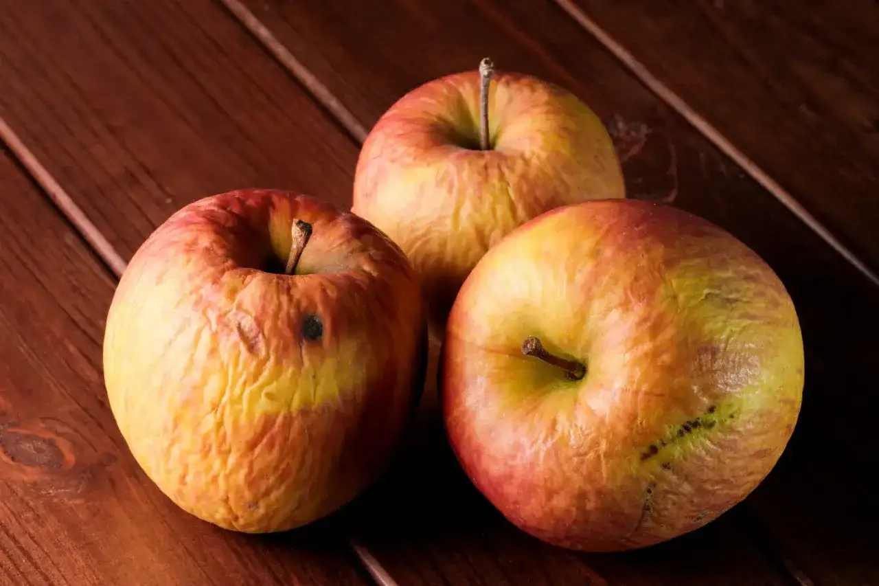 Trois pommes l&eacute;g&egrave;rement rid&eacute;es sur une table en bois. Parfaites pour savoir que faire avec des pommes ab&icirc;m&eacute;es : compote, tarte ou jus !