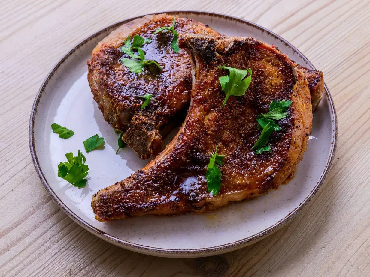 Deux belles côtes de porcelet grillées, nappées d'une sauce brillante et garnies de persil frais, sur une assiette.