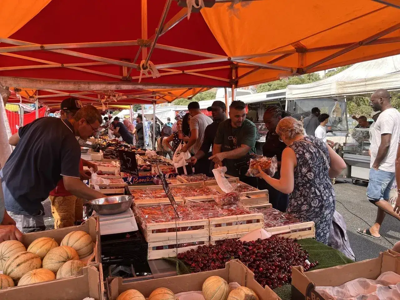 Un marché animé avec des étals débordant de fruits et légumes d'été. Des clients choisissent des cerises et des melons.