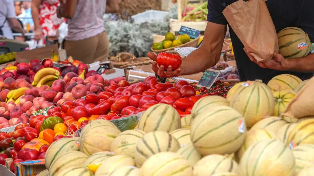 Un étal de marché débordant de fruits et légumes de saison, parfaits pour les salades de juin. Tomates, pêches, bananes et melons sont à l'honneur.
