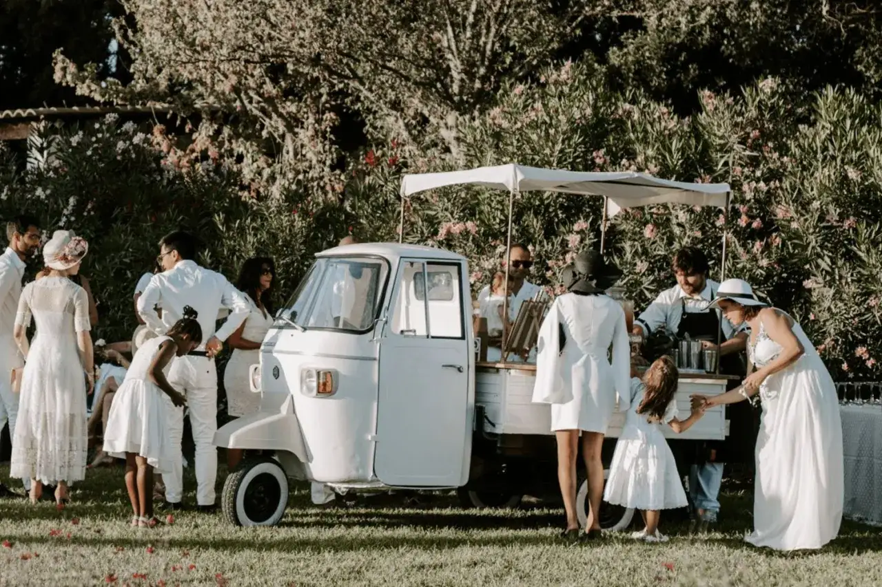 Un charmant triporteur blanc sert de bar à cocktail pour un mariage, avec des invités en tenues claires profitant de la fête en plein air.