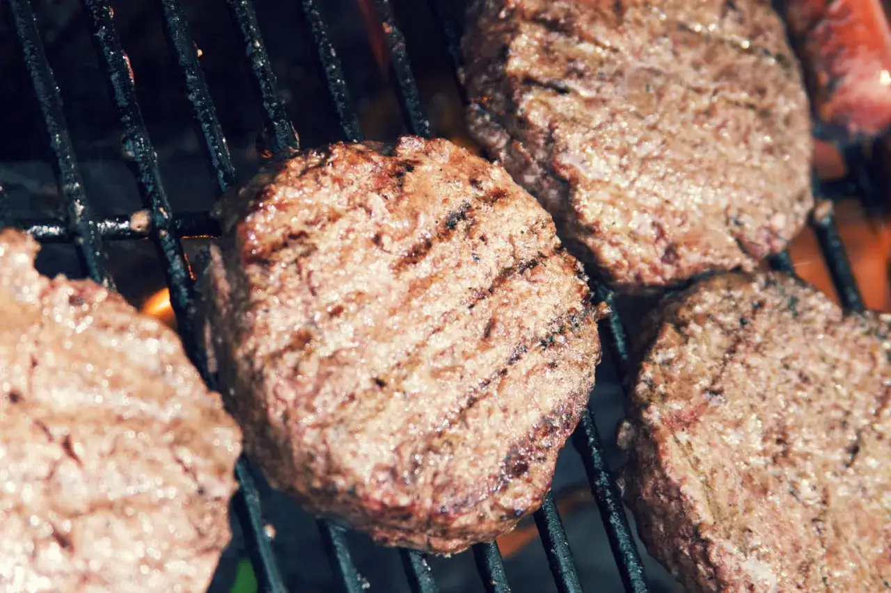 Quatre steaks hachés au barbecue grillent sur une grille, avec des flammes visibles en dessous.