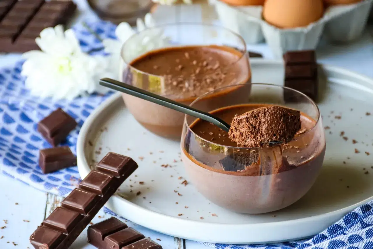 Deux coupes de mousse au chocolat skyr, garnies de cacao, avec des carr&eacute;s de chocolat et des fleurs blanches en arri&egrave;re-plan.