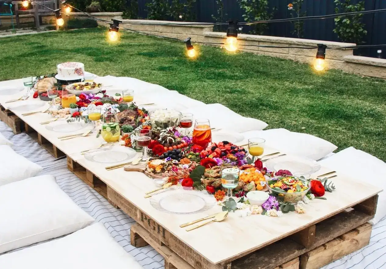 Un magnifique buffet champêtre pour un anniversaire, avec une table basse garnie de fruits, de viandes, de gâteaux et de fleurs, le tout sous des guirlandes lumineuses.