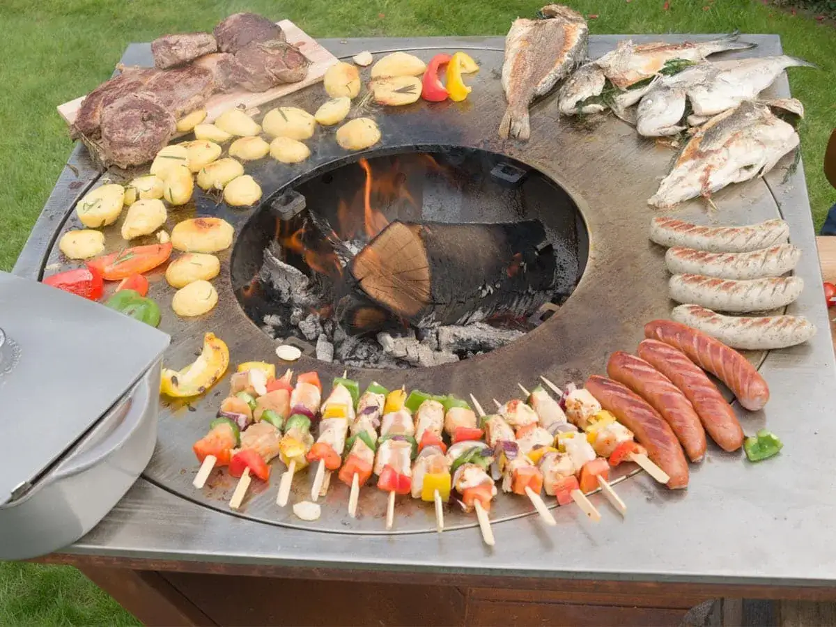 Un brasero barbecue fait maison, prêt pour un festin : viandes, poissons, saucisses, brochettes et légumes grillent sur la plaque chauffante.
