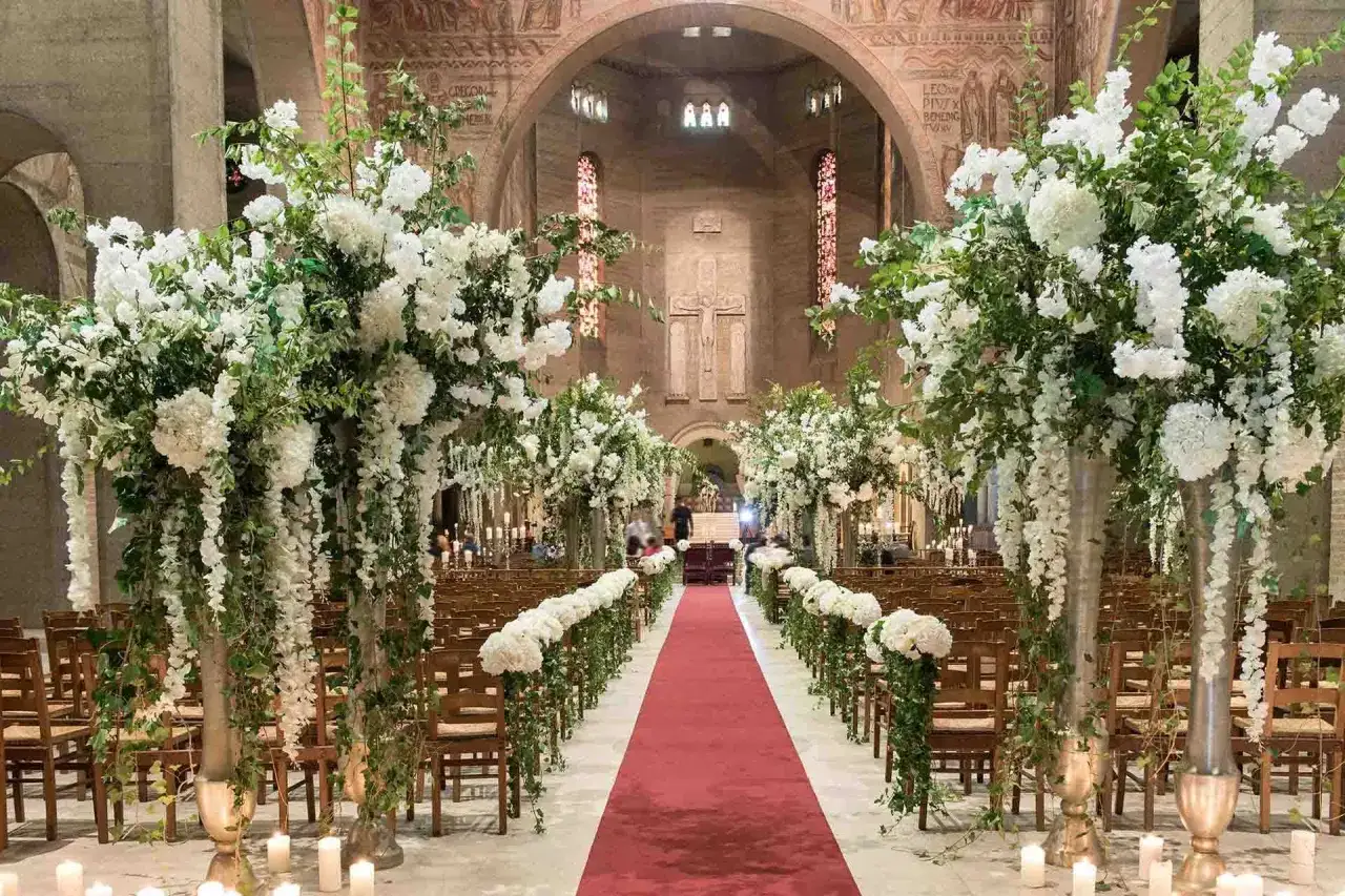 Décoration florale somptueuse pour un mariage à l'église. Le déroulement du mariage est marqué par un tapis rouge, des chaises en bois et des arrangements de fleurs blanches.