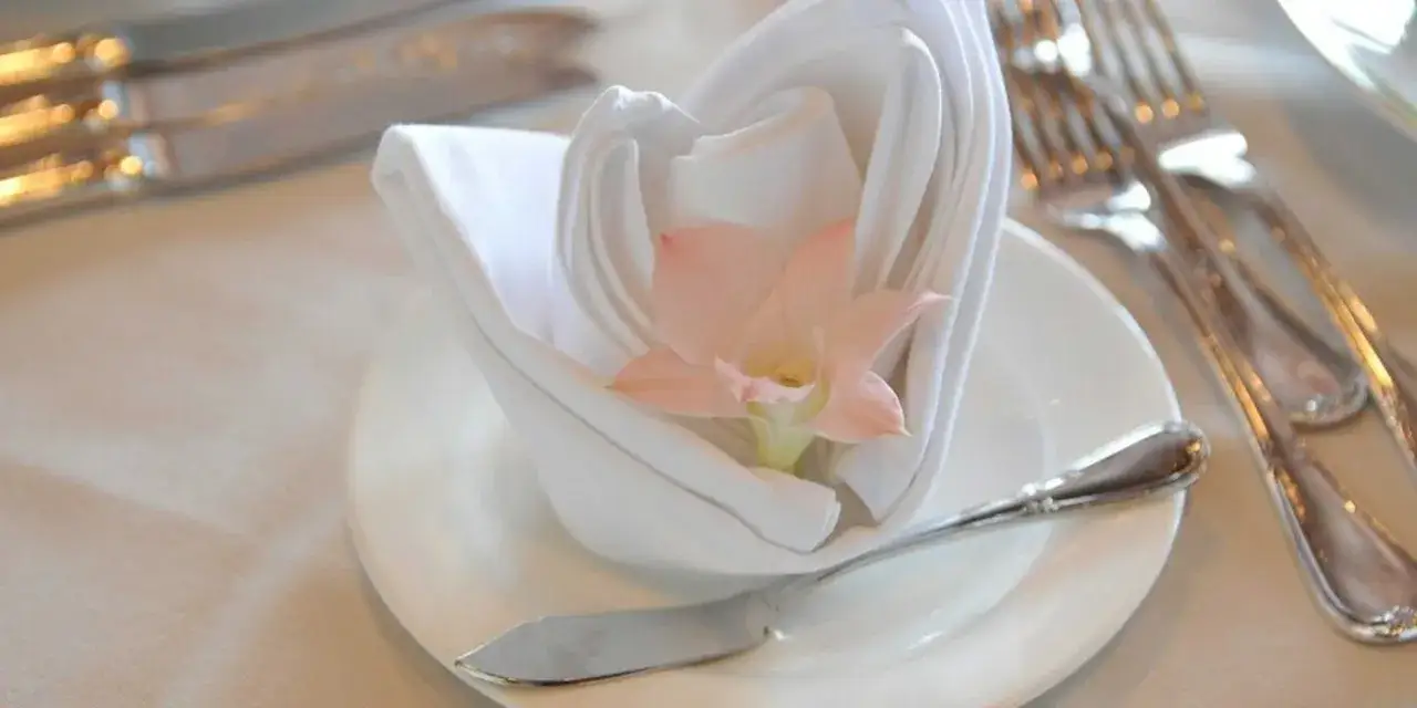 Un élégant pliage serviette mariage avec une fleur rose pâle, sur une assiette blanche, entouré de couverts argentés.