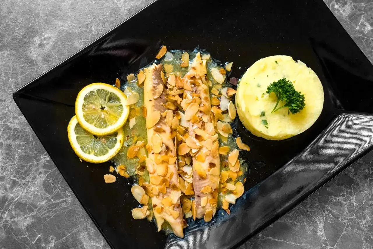 Délicieuse truite aux amandes, servie avec une purée de pommes de terre et des rondelles de citron sur une assiette noire.