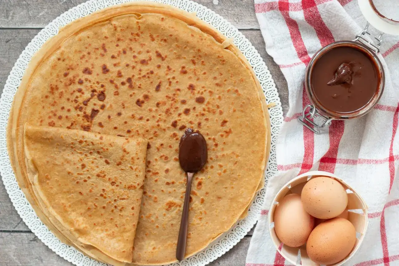 Pile de crêpes sans lactose, garnies d'une cuillère de pâte à tartiner au chocolat. Des œufs et un pot de pâte à tartiner sont à côté.