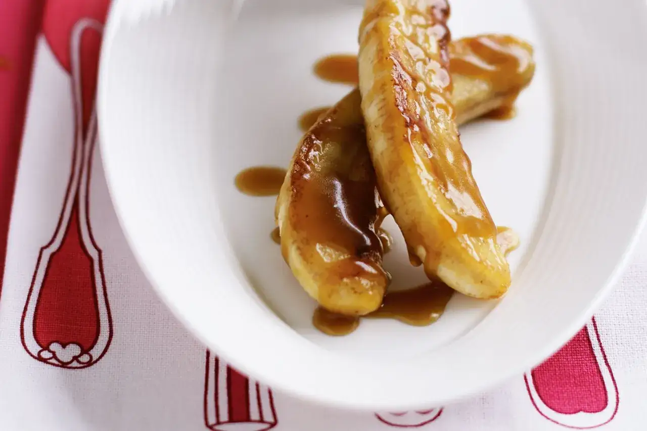 Deux bananes trop mûres, caramélisées à la poêle, nappées d'une sauce dorée, sur une assiette blanche.