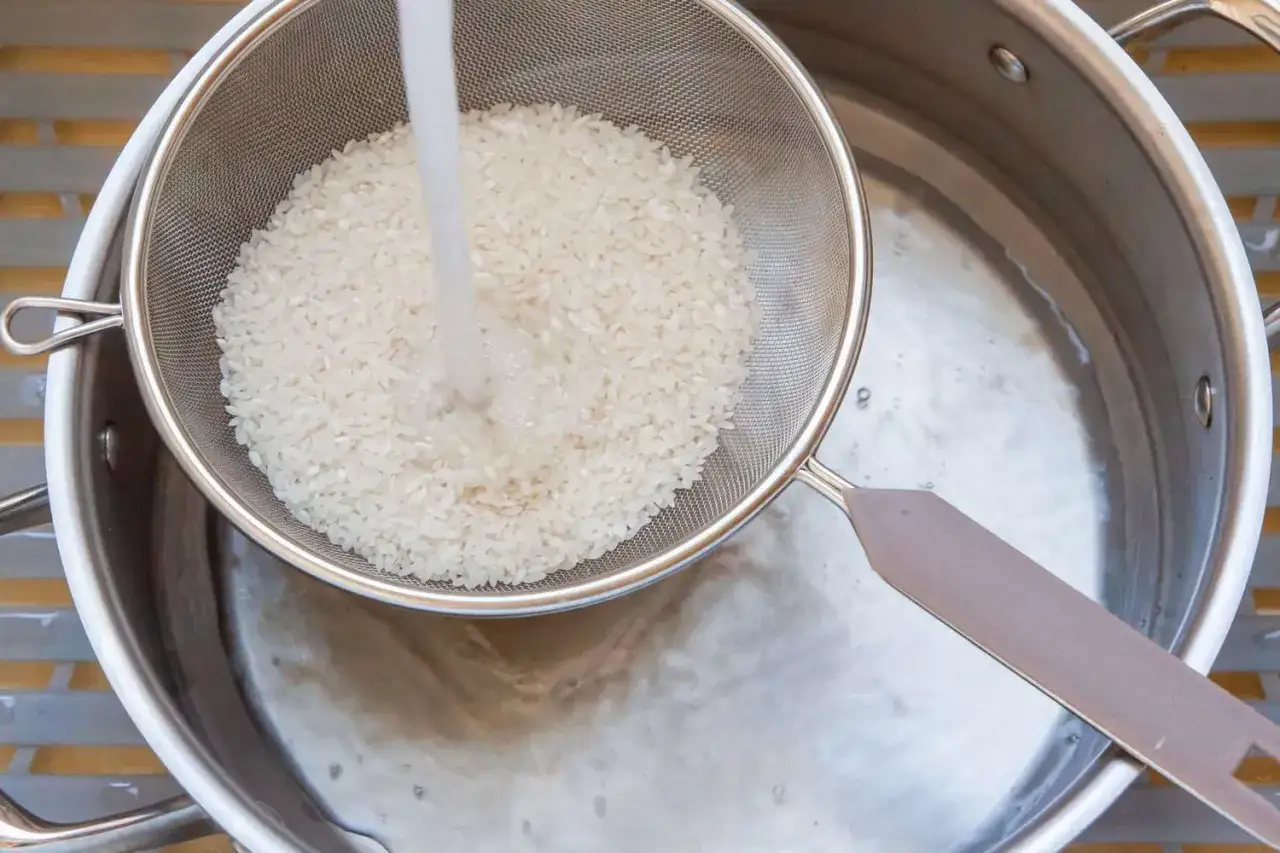Un jet d'eau claire coule sur du riz dans une passoire, expliquant pourquoi rincer le riz avant cuisson pour un résultat parfait.