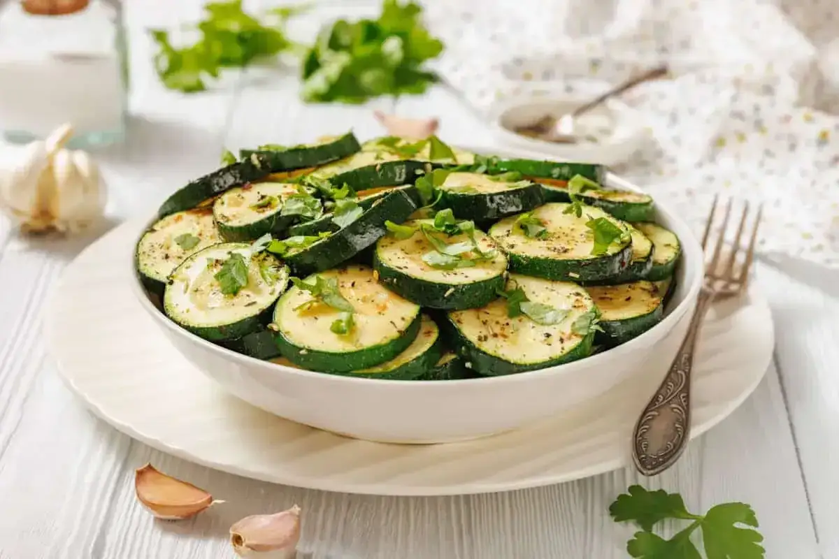 Tranches de courgettes grillées, garnies d'herbes fraîches, un plat d'été raffiné, servi dans un bol blanc avec de l'ail et du persil à proximité.